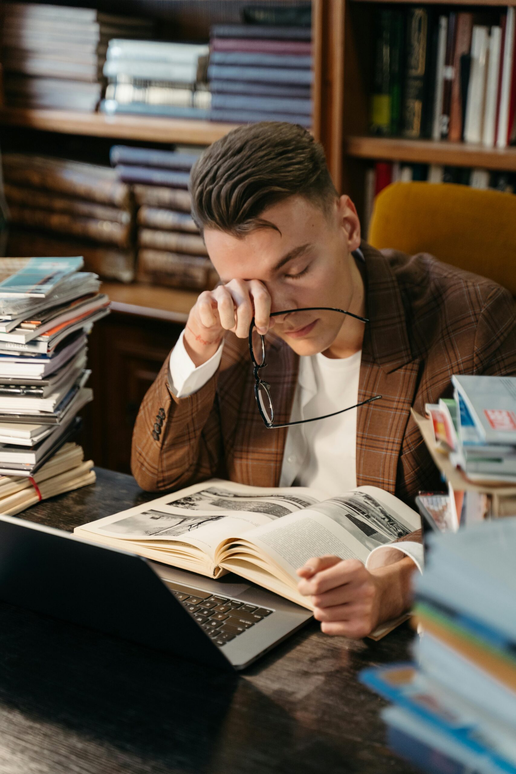 A tired student struggling to study in a library with piles of books and a laptop on the desk.